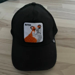Svart truckerkeps med bulldogpatch - Svart truckerkeps med mesh på sidorna och justerbar snapback. Framtill sitter en broderad patch med en orange och vit bulldog och texten 'BUTCH'. Kepsen har böjd skärm och klassisk form, perfekt för en avslappnad streetstil.
