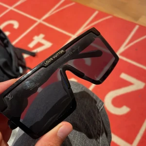 Svarta Louîs vuitŧon solglasögon - Säljer ett par svarta Louịs solglasögon med bred, rektangulär form och coola LV-detaljer i guld på bågarna. Glasögonen har en chunky design och mörka linser, perfekt för dig som vill sticka ut med en lyxig och modern look.