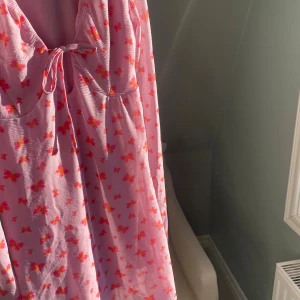 Rosa NA-KD klänning med fjärilar - Supersöt rosa klänning från NA-KD med lång ärm och v-ringning. Klänningen har ett gulligt fjärilsmönster i rosa och orange samt knyt framtill. Tunt, lätt material som känns luftigt och perfekt för varmare dagar.