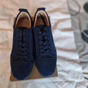 Mörkblå sneakers Christian Louboutin - Snygga mörkblå sneakers från Christian Louboutin i mocka med klassisk snörning och rund tå. Skorna har en ikonisk röd sula med Louboutin-signatur och broderad logga på hälen. Perfekt för dig som vill sticka ut med exklusiv stil.