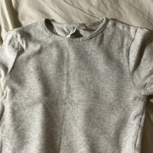 Ljusgrå ribbad topp från H&M Divided - Säljer en ljusgrå ribbad topp från H&M Divided Basic. Toppen har rund halsringning och korta ärmar, perfekt för en chill och enkel stil. Materialet är mjukt och stretchigt, vilket gör den skön att bära. Passar dig som gillar minimalistiska och cleana plagg.