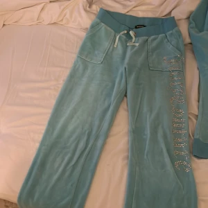 Juicy Couture blå set - Blåa mjukisbyxor från Juicy Couture i mjuk velour med bred resår i midjan och snörning. Byxorna har raka ben, fickor fram och bak samt glittrig Juicy Couture-text i strass längs ena benet. Perfekta för en chill och trendig look. Dem är väldigt små i storleken, skulle säga att det mer är som S/M.