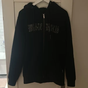 Svart hoodie från Wasted Paris med strass - Svart hoodie från Wasted Paris med strassdetaljer på bröstet och dragkedja framtill. Tröjan har huva med snörning och långa ärmar. Perfekt för dig som gillar streetwear och vill sticka ut med coola detaljer.