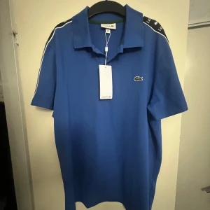 Blå pikétröja från Lacoste  - Snygg blå pikétröja från Lacoste i regular fit. Tröjan har klassisk krage, korta ärmar och svarta logoband med vita Lacoste-tryck längs axlarna. Ikoniska krokodilloggan på bröstet. Perfekt för en sportig och stilren look.