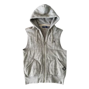 Grå hoodie-väst från Polo Ralph Lauren - Grå quiltad väst med huva och snörning från Polo Ralph Lauren. Västen har dragkedja framtill, två fickor och den klassiska broderade Polo-loggan på bröstet. Tillverkad i mjuk bomullsmix, perfekt för lager-på-lager och chill stil. Väldigt bra skick, liten fläck på ena fickan (bild 5) men inget man märker av när man använder den.