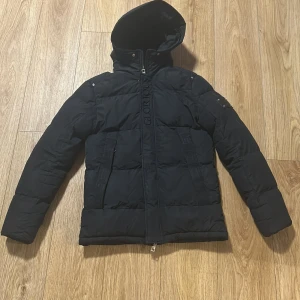 Svart pufferjacka Glorious Gangsta - Svart pufferjacka från Glorious Gangsta med huva och dragkedja. Jackan har en stor logga på ryggen och en snygg läderpatch med metallnitar på ärmen. Fylld för extra värme och har en clean, urban vibe. Perfekt för kalla dagar.