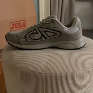 Beige Dior sneakers med CD-logga - Snygga beige sneakers från Dior med tydlig CD-logga på sidan och hälen. Skorna har en rund tå, platt sula och är tillverkade i en mix av skinn och mesh för en modern look. Svarta detaljer och snörning framtill. Perfekta för dig som gillar exklusiva streetwear-vibbar.