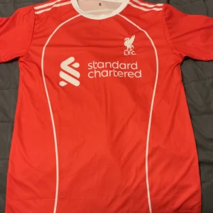 Liverpool Salah matchtröja röd S - Röd Liverpool FC fotbollströja med vita detaljer, tryck av 'M. Salah' och nummer 11 på ryggen. Framsidan har klubbmärke och sponsorlogga. Klassisk rund hals och korta ärmar. Tillverkad i lätt och ventilerande material, perfekt för fotboll eller support.