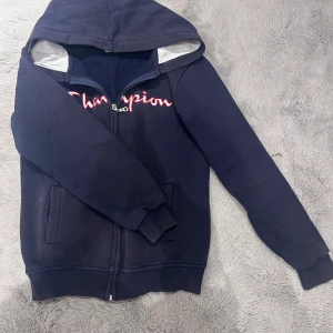 Mörkblå hoodie från Champion - Säljer en mörkblå hoodie från Champion med dragkedja och huva. Har lite målar färg på ärmen som jag tror kommer försvinna efter några tvättar eftersom det var mer tidigare. 