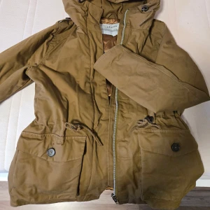 Vinterjacka från Elvine med huva - Snygg vinterjacka från Elvine med huva, dragkedja och stora fickor med knappar. Jackan har quiltat foder i satinliknande material och dragsko i midjan för en schysst passform. Perfekt för kyligare dagar och enkel att styla till det mesta.