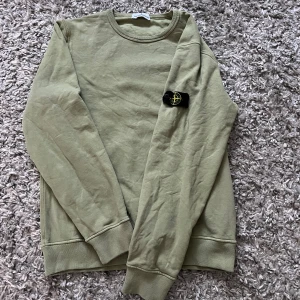 Olivgrön Stone Island sweatshirt - Snygg olivgrön sweatshirt från Stone Island med klassisk rund hals och ikonisk patch på ärmen. Tröjan är i mjuk bomull och har ribbade muddar vid ärmslut och nederkant. Perfekt för dig som gillar streetwear och vill ha en clean look.