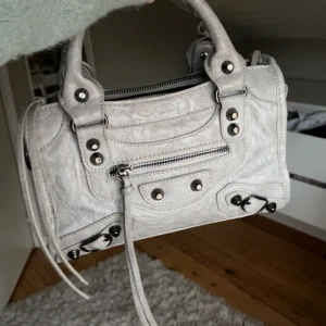 Grå handväska  - Säljer en snygg ljus grå handväska i skinn med coola metalldetaljer och dragkedja. Det tillkommer även ett långt band som man ser på andra bilden 