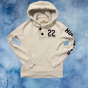 Vit Vintage Hollister Hoodie Med Knappar - Sällsynt vintage Vit hoodie från Hollister med broderat '22 Surf Team' på bröstet och stort Hollister-tryck längs ena ärmen. Tröjan har huva med snörning, knappar vid halsen och en klassisk känguruficka. (Små svarta prickar finns på hoodien men syns knappast då vintage hoodies har sliten/använd design). Perfekt för dig med avslappnad stil! 