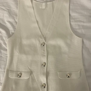 Vit väst med silvriga knappar från H&M - Stilren vit väst från H&M med v-ringning och silvriga knappar framtill. Västen har två fickor med lock och knappdetalj. 