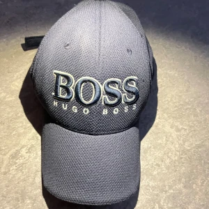 Mörkblå keps från Hugo Boss - Snygg mörkblå keps från Hugo Boss med stor broderad logga framtill. Kepsen har justerbar rem baktill med metallspänne och insida i limegrönt och beige. Tillverkad i ett grovt vävt material som ger en sportig känsla.