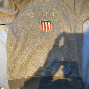 Grå hoodie från GANT med logga - Säljer en grå hoodie från GANT med broderad logga och sköld på bröstet. Tröjan har huva, magficka och långa ärmar. Perfekt för dig som gillar klassisk och sportig stil.