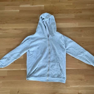 Ljusgrå hoodie med dragkedja - Mysig ljusgrå hoodie med dragkedja framtill och stor huva. Tröjan har två fickor på magen och är tillverkad i mjuk bomullsblandning. Perfekt för chill dagar eller när du vill ha något bekvämt och enkelt.