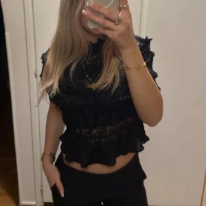 Svart volangblus i spets - Säljer en svart blus med korta ärmar och hög hals. Blusen är täckt av volanger och har ett genomgående spetsmaterial som ger en cool och romantisk vibe. Perfekt för dig som gillar detaljer och vill sticka ut lite extra.