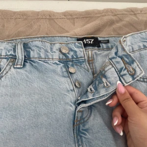 Ljusblå jeanskjol från 157 - Snygg ljusblå jeanskjol från 157 med klassisk femficksdesign och knappar framtill. Kjolen har bälteshällor och är tillverkad i bomull. Perfekt för dig som gillar en enkel och stilren denimlook.