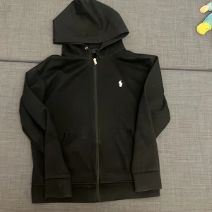 Svart hoodie från Polo Ralph Lauren - Svart hoodie från Polo Ralph Lauren med klassisk vit broderad logga på bröstet. Tröjan har huva, dragkedja framtill och två fickor. Materialet är mjukt och känns som bomull, perfekt för chill dagar. Snygg och enkel design som funkar till allt.