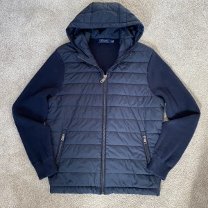 Polo Ralph Lauren Hybrid Knit Cardigan - Säljer en mörkblå jacka från Polo Ralph Lauren med huva och dragkedja framtill. Jackan har quiltad framsida, stickade ärmar och två fickor med dragkedja. Perfekt för kyliga dagar och riktigt snygg till jeans eller joggers.