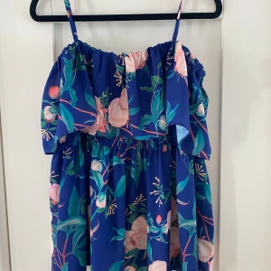 Blommig blått linne med volang - Säljer en superfin blått linne med justerbara smala axelband och volang upptill. Linnet har ett färgstarkt blommigt mönster i rosa och grönt, samt smock i midjan för en skön passform. Perfekt för sommarens alla tillfällen! Mått Över bröstet: ca 41 cm Längd från axeln och ner: ca 61-68 cm  100% polyester   Kommer från ett djur och rökfritt hem. 