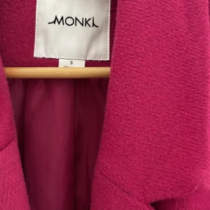 Ceriserosa kappa från Monki - Säljer en ceriserosa kappa från Monki med dubbelknäppning och stora fickor framtill. Kappan har klassisk krage och är helfodrad, perfekt för dig som vill sticka ut i höst och vinter. Materialet är mjukt och känns lyxigt.Den är lite nobbig på vissa delar och syns knappt annars är det inget fel på den. 