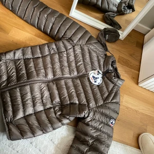 Brun dunjacka JOTT - Snygg brun dunjacka från JOTT  med horisontella sömmar och dragkedja framtill. Jackan har huva och klassisk logga på ärmen. Perfekt för kyliga dagar och lätt att packa ner i medföljande påse. Aldrig använd 