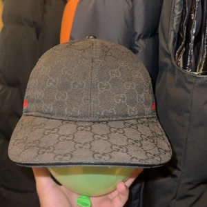 Svart keps från Gucci med GG-mönster - Säljer en svart keps från Gucci med klassiskt GG-monogram över hela kepsen. Sidorna har gröna och röda band som ger den en lyxig touch. Kepsen är i bomull och har justerbar rem bak för perfekt passform. Perfekt accessoar för att lyfta din outfit.
