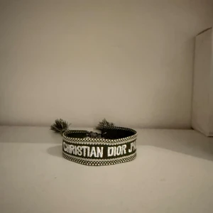 Christian Dior J'Adior armband - Snyggt armband från Christian Dior med texten 'J'Adior' broderad i vitt på en mörkgrön bakgrund. Armbandet har justerbar knytning med tofsar i ändarna och detaljerad brodyr längs kanterna. Perfekt statement-piece för att lyfta din outfit.