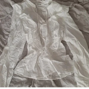 Vit volangblus från Kappahl, stl S - Supersöt vit blus från Kappahl i storlek S. Blusen har långa ärmar, volangdetaljer längs halsringningen och framtill, samt en lätt skrynklig look. Perfekt för dig som gillar romantiska och feminina detaljer.