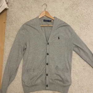Grå kofta från Polo Ralph Lauren - Snygg grå kofta från Polo Ralph Lauren med klassisk v-ringning och knappar framtill. Tröjan har två fickor och den ikoniska broderade loggan på bröstet. Tillverkad i mjuk bomull och har långa ärmar. Koftan passar inte mig därav ligger den ute till försäljning. Om intresserad är det bara att skriva:) Pris kan diskuteras😊
