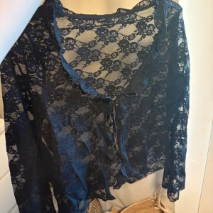 Svart spetsig topp - Säljer en svart bolerotopp i genombruten spets med blommigt mönster. Toppen har långa ärmar och en söt snörning framtill. Perfekt för att lägga över en linne eller klänning för en extra touch. Supersnygg och trendig detalj till din outfit!