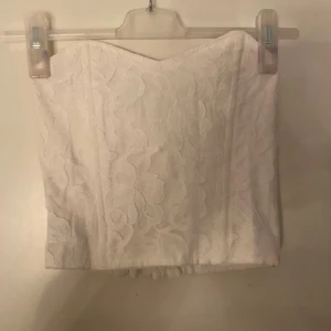 Vit spets tubtopp med smock - Säljer en vit tubtopp med blommig spets och smockad rygg. Toppen är axelbandslös och har en snygg bandeauform. Materialet är mjukt och har en lätt struktur med blommönster. Perfekt för sommarens alla tillfällen, superfin att styla med jeans eller kjol. Helt oanvänd!super snygg.🤩 