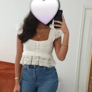 Vit spetsig blus med volang från zara  - Säljer en superfin vit blus med spetsdetaljer och volang nedtill. Toppen har korta ärmar med spets och en fyrkantig ringning. Materialet är bomull med broderade mönster och genombruten spets. Perfekt för dig som gillar romantisk och feminin stil.