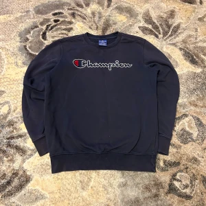 Marin blå Champion sweatshirt i bomull - Marin blå sweatshirt från Champion med broderad logga i vitt och rött på bröstet. Klassisk rund halsringning och långa ärmar. Mjuk insida och ribbade muddar vid ärmslut och nederkant. Perfekt för en chill stil.