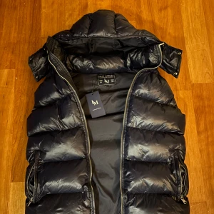 Mörk blå väst från Leon Marcel - Osndvänd!! Snygg svart dunväst från Leon Marcel med avtagbar huva och två dragkedjeförsedda fickor. Västen är quiltad och har en rak passform med blanka paneler. Perfekt för lager-på-lager och ger en cool vibe till din outfit.