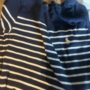 Randig blå tröja från Polo Ralph Lauren - Snygg marinblå tröja från Polo Ralph Lauren i storlek S med vita horisontella ränder och klassisk broderad logga på bröstet. Tröjan har lång ärm och är gjord i mjuk bomull, perfekt för en chill och stilren look.