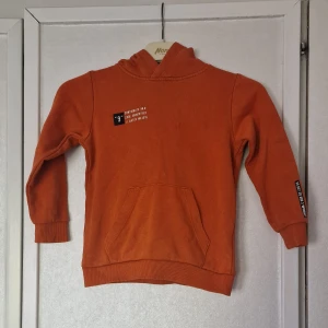 Orange hoodie från WOXO 720° - Orange hoodie från WOXO 720° med tryckdetaljer på bröstet och ärmen. Tröjan har huva, magficka och ribbade muddar vid ärmslut och nederkant. Färgen har blivit något tvättpåverkad och är inte längre lika lysande orange som den var när den köptes och det är några fläckar som inte har gått bort i vanligt tvättprogram. Ingen tröja att ha när man ska klä upp sig men däremot perfekt när man leker eller är på förskolan.
