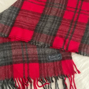 Rutig röd och grå halsduk från Burberrys - Klassisk halsduk från Burberrys i rött och grått rutigt mönster med fransar i ändarna. Tillverkad i 100% lammull, mjuk och varm. Perfekt accessoar för att lyfta din vinterstil med en ikonisk brittisk touch.