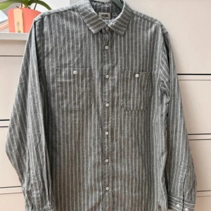 Vans skjorta  - Snygg grå skjorta med vita vertikala ränder från Vans. Klassisk krage, två bröstfickor med knappar och diskret Vans-märke vid nederkanten. Långärmad och tillverkad i mjuk bomull, perfekt för en chill och stilren look.