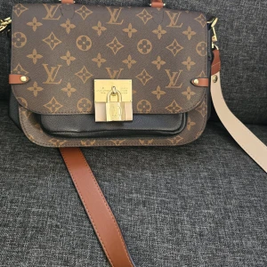 Väska från välkänt märke  - Snygg axelväska från välkänt märke med klassiskt monogrammönster i brunt och beige. Väskan har guldfärgade metalldetaljer, ett lås framtill och bred justerbar axelrem i brunt och beige. Tillverkad i skinn med strukturerad form och flera fack.