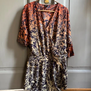 Mönstrad omlottklänning  - Klänning med abstrakt mönster i orange, beige och mörkblått. Klänningen har v-ringning, resår i midjan trekvartsärmar. 