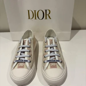 Beige och vita sneakers från Dior - Snygga sneakers från Dior i beige och vitt med diskret mönster och tydlig Christian Dior-logga på snörningen. Skorna har vit platt sula med svart stjärndetalj undertill och är tillverkade i textil med skinnliknande detaljer. Perfekta för dig som vill ha en exklusiv och trendig look . Skorna är endast använda 1 gång och har därefter stått i garderoben.