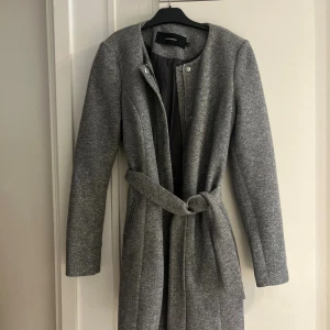 Grå kappa från Vero Moda med bälte - Stilren grå kappa från Vero Moda med knytbälte i midjan och diskreta knappar. Jackan har rundad halsringning, långa ärmar och är tillverkad i ett mjukt material som känns både klassiskt och modernt.