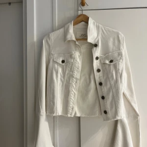 Vit croppad jeansjacka Viva Couture - Snygg vit croppad jeansjacka från Viva Couture med rå kant nertill och klassiska bröstfickor med knappar. Jackan har långa ärmar och metallknappar framtill. Perfekt för dig som vill ha en fräsch och trendig look.