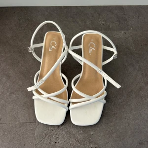 Vita klackar med låg klack från Clou - Snygga vita klackar från Clou med flera tunna remmar och justerbart spänne vid ankeln. Endast använd en gång.