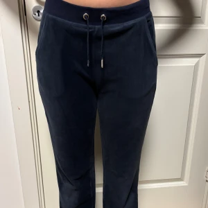 Juicy Couture svarta mjukisbyxor - Mörk blåa mjukisbyxor från Juicy Couture i mjuk velour med glittrande strasstenar som bildar loggan på ena benet och stor 'Juicy' text bak. Byxorna har snörning i midjan och två fickor fram. Perfekta för en chill och trendig look.