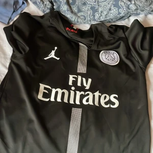 Svart PSG x Jordan fotbollströja S - Säljer en svart PSG x Jordan fotbollströja i storlek S. Tröjan har korta ärmar, vit Paris Saint-Germain-logga och Fly Emirates-tryck på bröstet. Materialet är lätt och andas, perfekt för match eller träning. Snyggt vertikalt randigt mönster framtill.