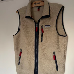 Patagonia Fleeceväst - Säljer en beige fleeceväst från Patagonia i storlek S. Västen har marinblå detaljer, tre fickor med dragkedja och röda zip-pullers. Mjuk och fluffig känsla, perfekt med hög krage och snygg kontrastkant runt ärmhålen. Lite smutsig på magen men syns inte vid användning!